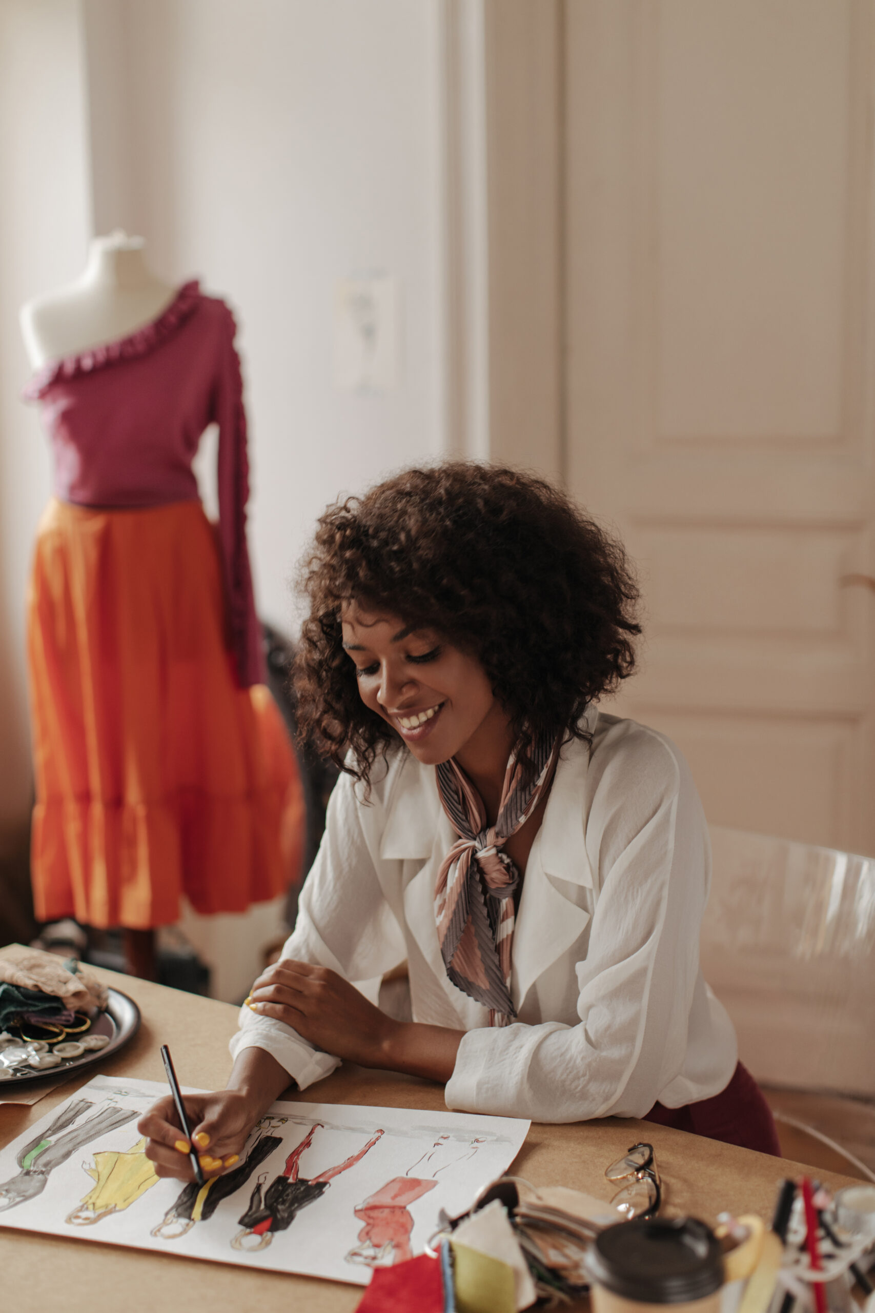 cheerful young dark skinned woman in white trendy blouse draws clothes samples. attractive fashion